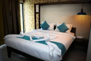 Neatly made bed with teal accents and towel swans in a floral room, inviting North Goa accommodation near Anjuna Beach.